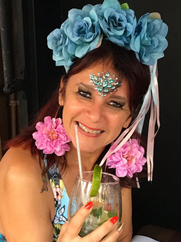 Andrea Magalhães usa tiara e brinco com flores enquanto segura um copo de bebida